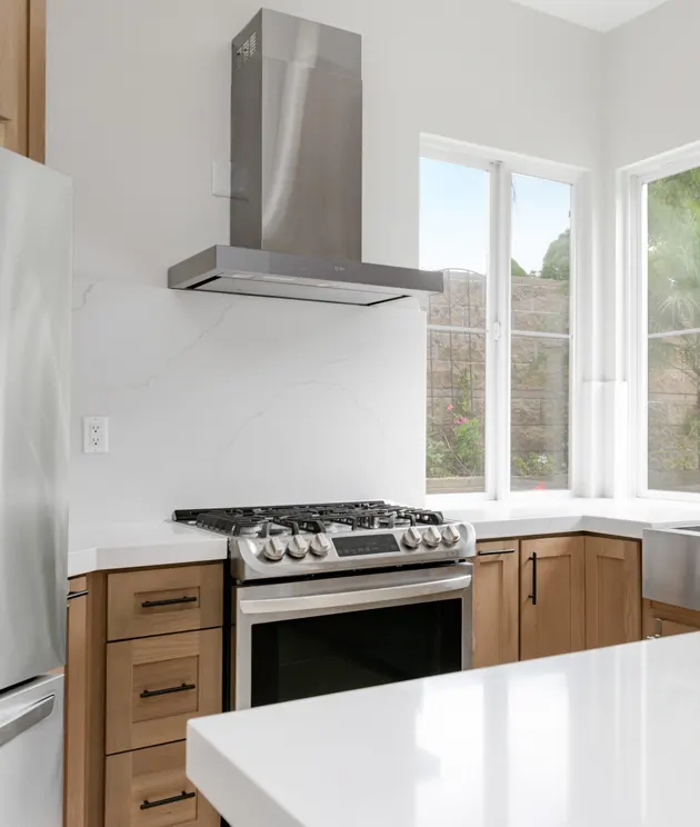 A modern kitchen featuring light wood cabinets with black hardware, a stainless steel slide-in gas range, and a sleek stainless steel range hood mounted on a white wall. White windows provide natural light above the countertop, and a white countertop or island is visible in the foreground. The kitchen has a clean, minimalist design with a neutral color palette of white, natural wood, and stainless steel.