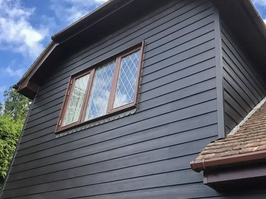 A dark gray weatherboard house exterior with horizontal cladding beneath a peaked roof. Two wooden-framed windows with diamond-pattern leaded glass are visible on the upper story. The image shows part of a tiled roof section on the right side, with trees and blue sky visible in the background.