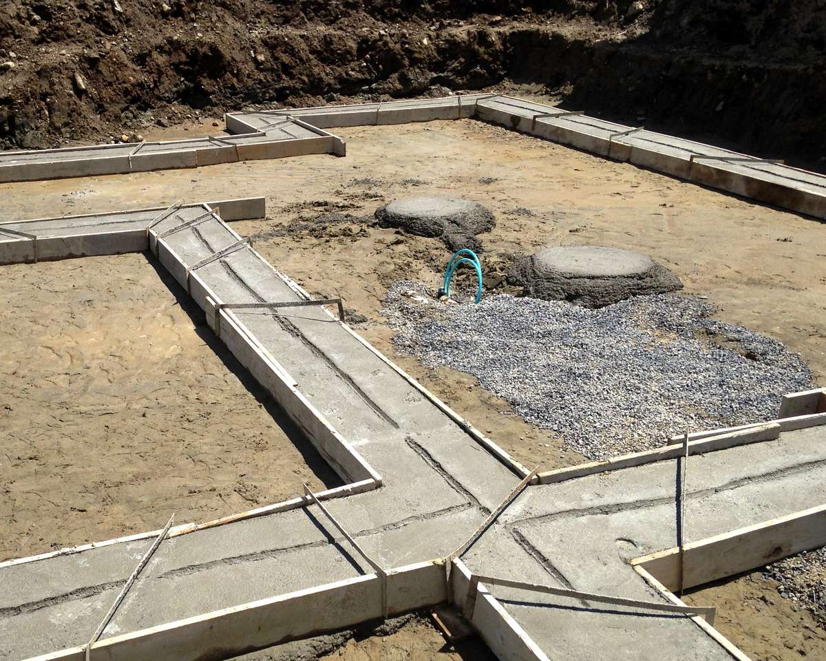 A concrete foundation under construction showing the initial stage of building work, with fresh concrete footings forming the perimeter and internal walls of a structure. The foundation is set in sandy excavated ground with dirt banks visible in the background. A section of gravel base is visible in the center, and a blue pipe or hose can be seen on the construction site.