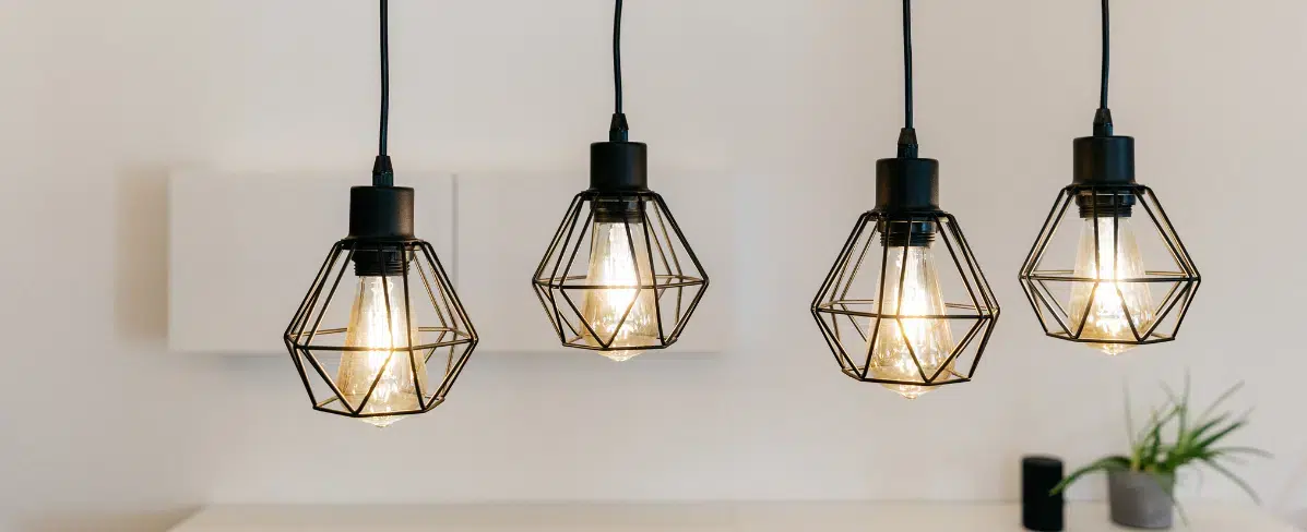 Four geometric pendant lights with black metal wire cages hang in a row against a white wall. Each cage-style fixture contains an illuminated Edison-style bulb creating a warm glow. The modern industrial lighting arrangement is complemented by minimalist decor including white frames and a small potted plant visible in the background.