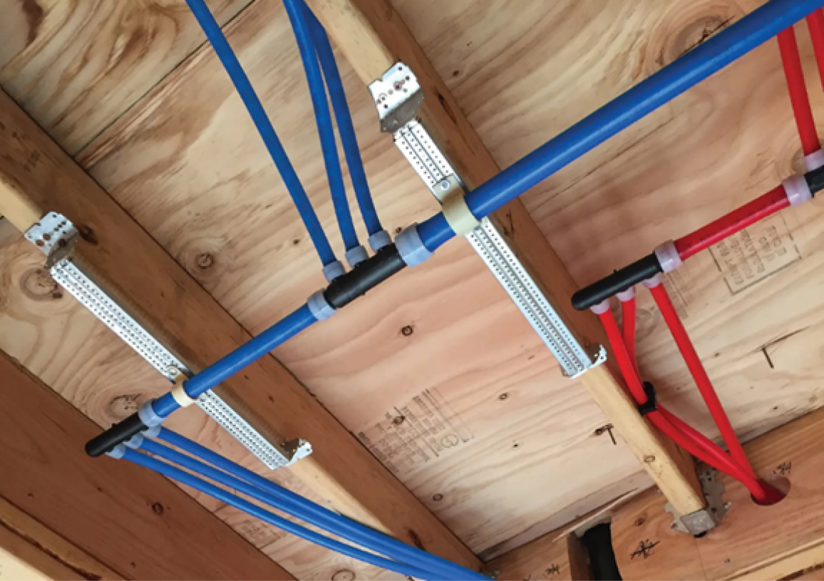 PEX plumbing installation in an unfinished ceiling showing blue and red PEX tubing running through wooden joists with white metal support brackets securing the pipes. The color-coded system uses blue for cold water lines and red for hot water lines, demonstrating modern residential plumbing methods in new construction or renovation work.