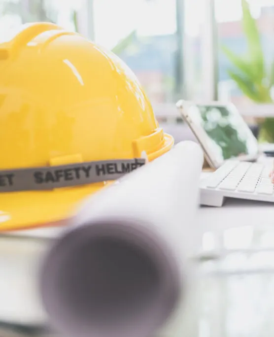 A bright yellow hard hat with "SAFETY HELMET" text sits on a white surface in the foreground, with architectural blueprints or construction plans visible but slightly out of focus in the background. The scene appears to be in an office or planning area with natural light coming through a window, suggesting a construction or engineering work environment.