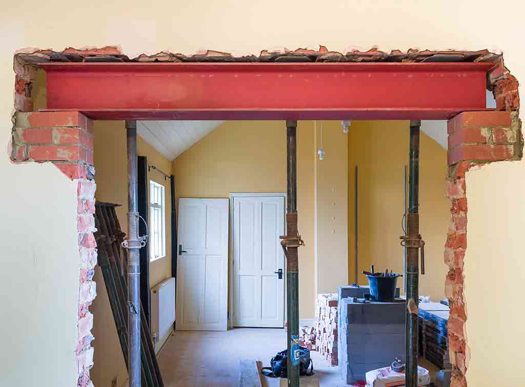 A residential interior under renovation showing a large opening in a brick wall with an exposed red steel beam spanning across the top. The wall has been partially demolished, revealing rough edges and exposed brickwork on both sides. Through the opening, a room with cream-colored walls, white doors, and construction materials scattered on the floor is visible.