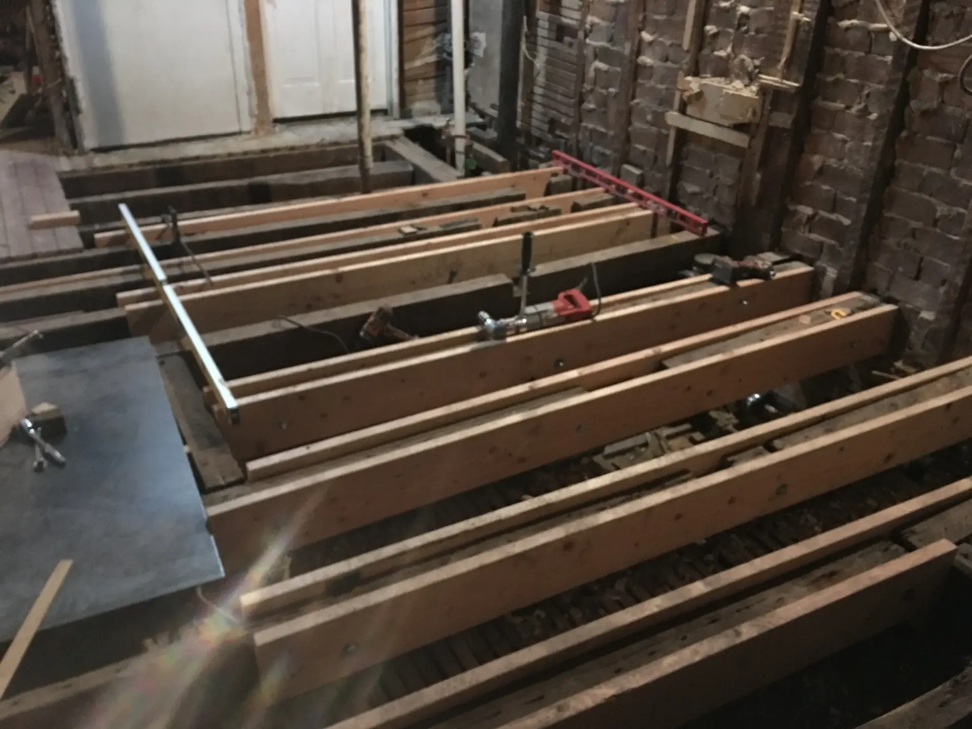 Construction in progress showing wooden floor joists installed across a room with exposed brick walls. The parallel timber beams create the structural framework for a new floor, with tools and construction materials visible among the framing. A doorway is visible in the background leading to another room.