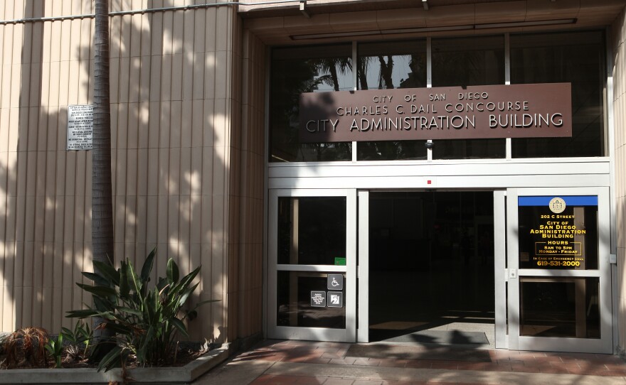 The entrance to the City of San Diego Charles C. Dail Concourse City Administration Building, featuring glass doors with metal frames and a brown sign above displaying the building name. A plaque is visible on the right side of the entrance, and palm trees cast dappled shadows on the beige corrugated exterior walls. The entrance has accessible features including what appears to be accessibility signage near the door.
