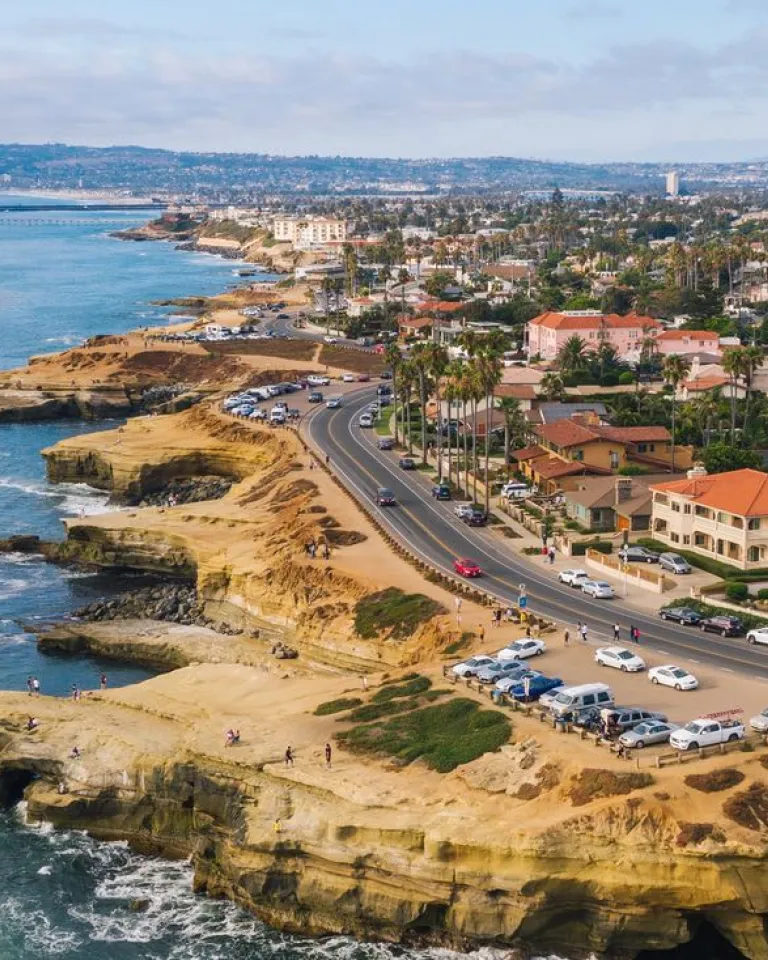 Aerial view of a San Diego coastal community featuring distinctive sandstone cliffs and rock formations along the Pacific Ocean shoreline. A curved highway runs parallel to the coast, lined with palm trees and residential buildings with Spanish-style terracotta roofs. The landscape shows both natural geological features and developed beachfront areas with parked vehicles and beach access points.