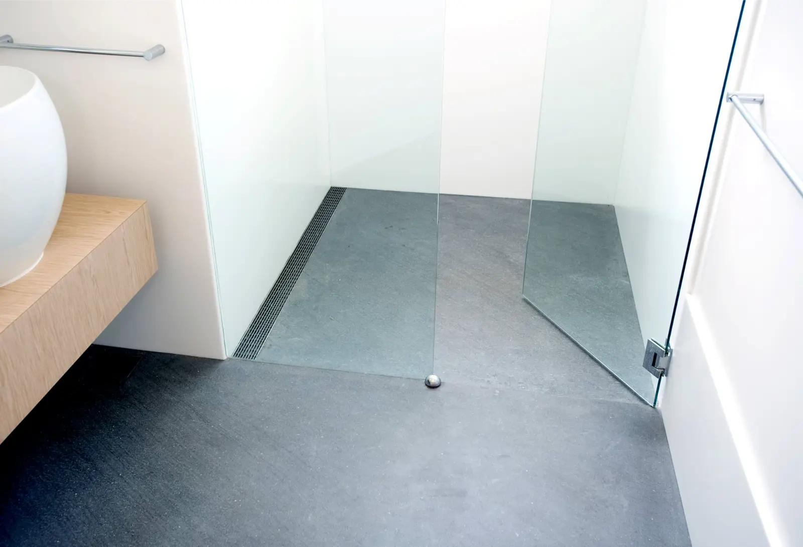 A modern minimalist bathroom featuring a curbless walk-in shower with gray slate tile flooring and a linear drain along one wall. The shower area is enclosed by a clear glass panel and includes white walls. A light wood floating vanity with a white vessel sink is visible on the left side of the image.