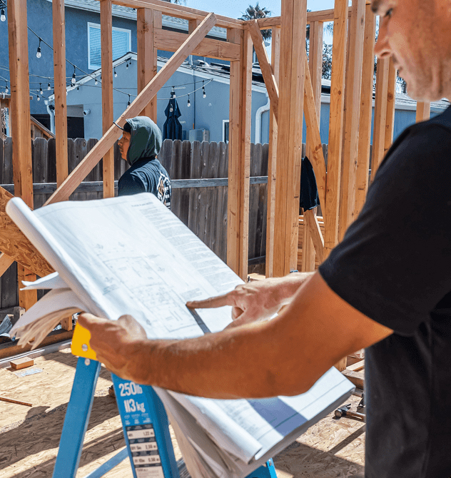 A construction worker in a black shirt reviews architectural blueprints at an active building site with wooden framing in progress. The blueprints are spread across a blue ladder while another worker in a hooded sweatshirt works on the frame structure in the background. A residential fence and neighboring buildings are visible behind the construction area.