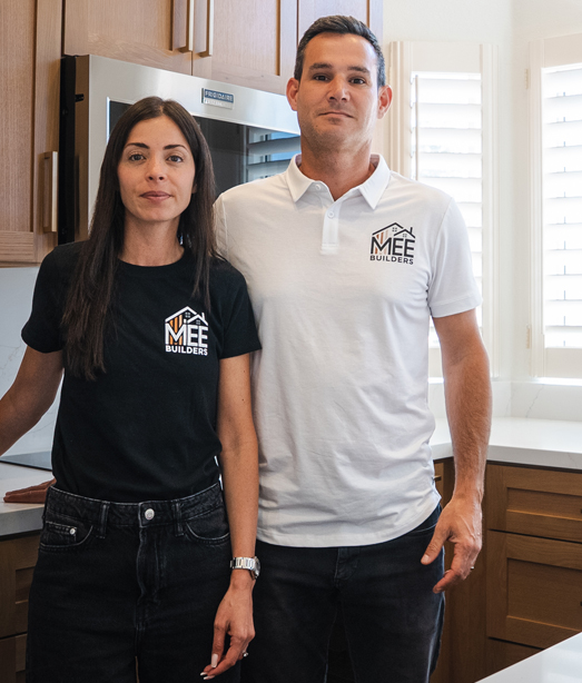 A man and woman stand together in a modern kitchen, both wearing matching polo shirts with an "MFF" logo. The woman wears a black shirt while the man wears white, and they appear to be business partners or team members. Behind them are white kitchen cabinets and dark wood lower cabinets in a contemporary residential setting.