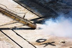 A pressure washer wand directs a powerful spray of water onto a concrete surface, creating a dramatic plume of mist and revealing a clean strip contrasting with the dirty surrounding area. The cleaning process demonstrates the stark difference between the weathered, stained concrete and the freshly cleaned section. The wooden deck boards are visible in the upper portion of the image, showing the residential setting of this outdoor cleaning project.