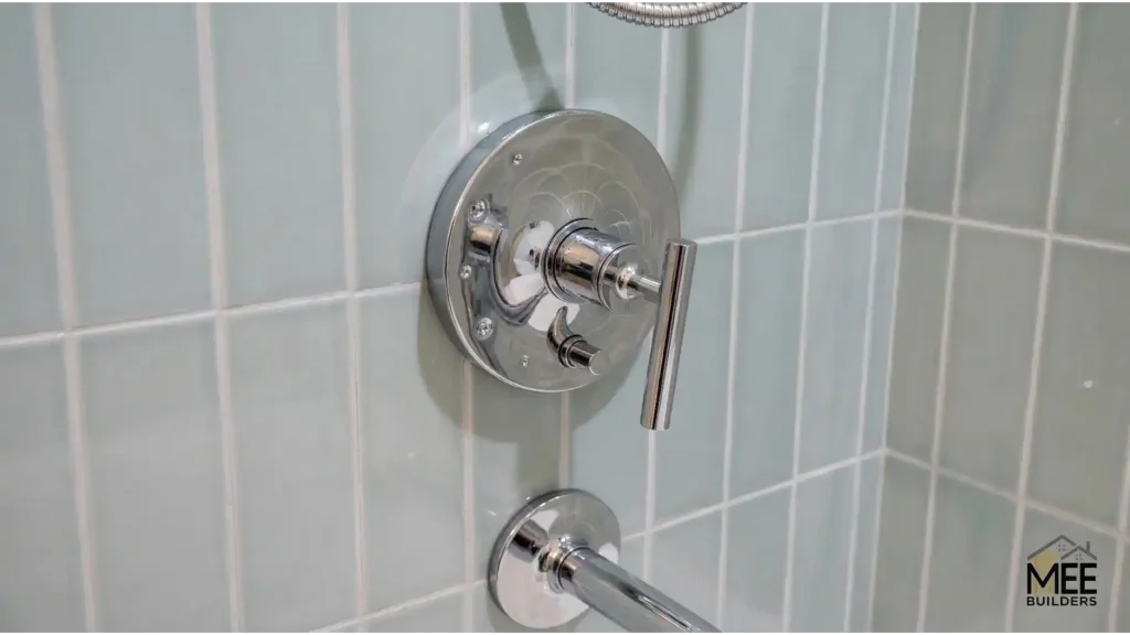 A chrome shower fixture mounted on light gray vertical subway tile with white grout. The fixture features a circular trim plate with a modern lever handle control and a secondary diverter below. The MEE Builders logo appears in the bottom right corner of the image.