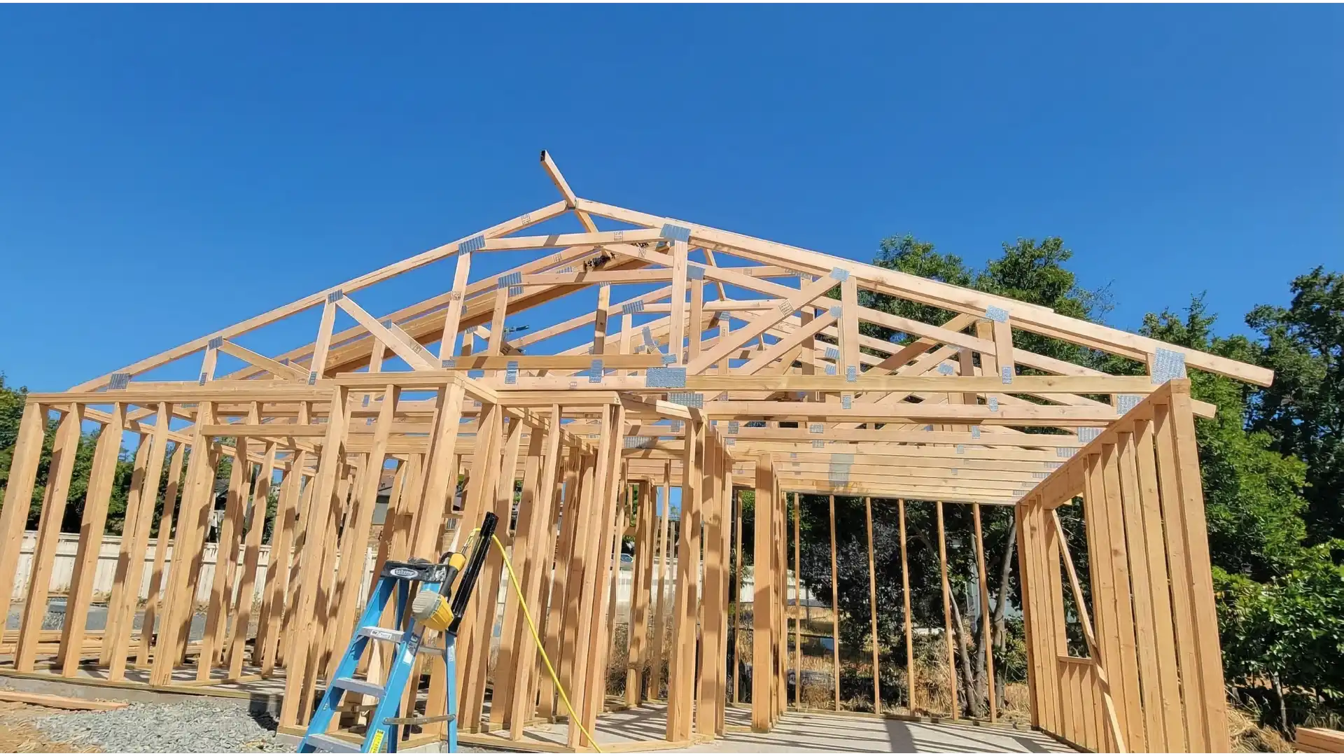 A residential building under construction showing the wooden frame structure with exposed roof trusses and wall studs. The skeleton of the house features light-colored lumber forming the structural framework against a clear blue sky, with a blue ladder positioned inside and trees visible in the background. The construction appears to be in the early framing stage before exterior sheathing and roofing materials are installed.