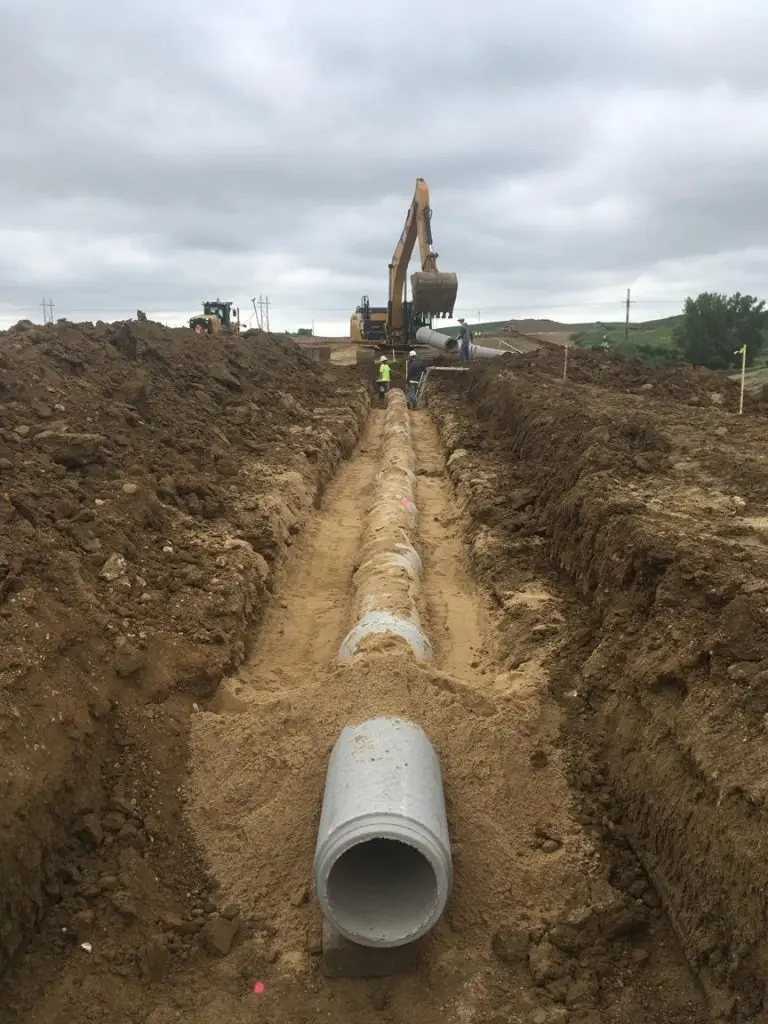 A construction site showing a long trench with large concrete pipes laid in sequence along sandy soil. An excavator is positioned in the background with a worker in safety vest standing nearby. The scene depicts pipeline infrastructure installation work under an overcast sky with rolling hills visible on the horizon.