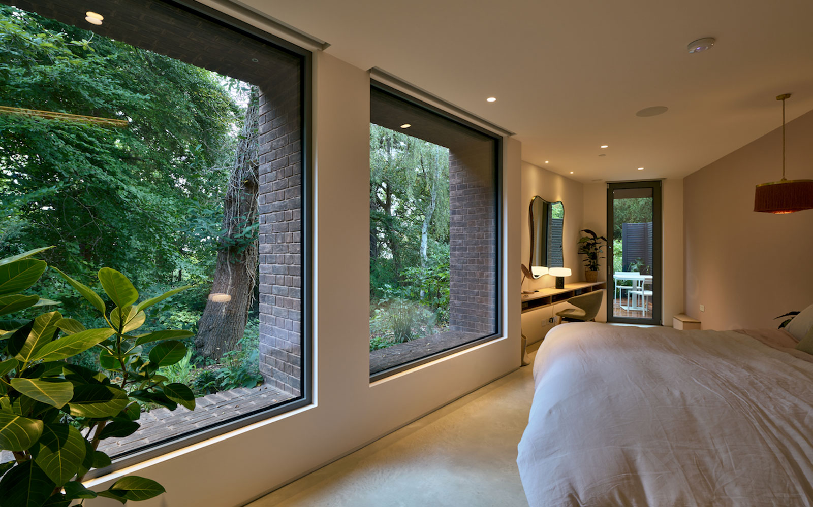 A modern minimalist bedroom featuring two large floor-to-ceiling windows with black frames overlooking lush green trees and foliage. The room has a clean white bed, recessed lighting, and a view through to an ensuite area with a vanity and access to an outdoor deck. An indoor potted plant with large green leaves is positioned near the left window, complementing the natural forest views.