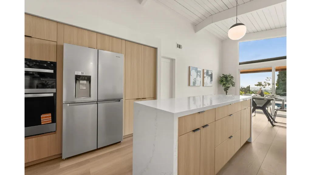 A modern kitchen featuring light wood cabinetry with stainless steel appliances, including built-in wall ovens and a four-door refrigerator. The space has a white waterfall-edge island, vaulted white wood ceiling with pendant lighting, and opens to an outdoor patio area through large glass doors. The minimalist Scandinavian-inspired design emphasizes clean lines and natural materials with light wood flooring throughout.