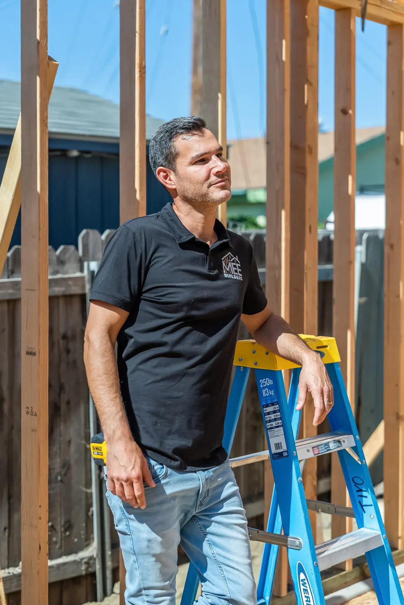 A builder wearing a black polo shirt with company branding stands at a construction site next to a blue and yellow stepladder. He is positioned among exposed wooden wall framing on a sunny day, looking off to the side in a thoughtful pose. The construction setting shows timber framework and a residential fence in the background.