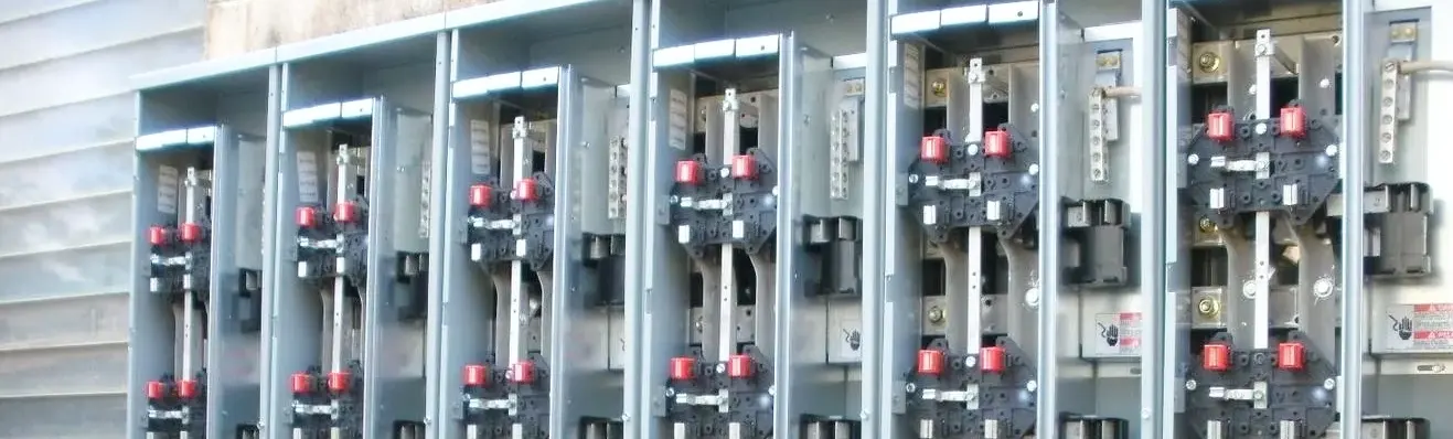 A row of electrical meter bases mounted on the exterior of a building, each containing dual red-topped meter sockets and dark circuit breaker panels. The metal enclosures are arranged in a horizontal line against light-colored siding, typical of multi-unit residential electrical service installations. Each unit features standardized electrical metering equipment with visible connection points and safety components.