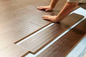 A person's hands installing laminate flooring planks on a light-colored underlayment. The worker is positioning a brown wood-textured plank next to already-laid flooring pieces, demonstrating the click-and-lock installation process. The image shows the detailed wood grain pattern on the laminate surface and the proper technique for fitting planks together during floor installation.