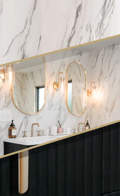 A modern bathroom vanity featuring a luxurious white marble wall with gray veining, two round gold-framed mirrors, and matching gold wall sconces. The vanity has white countertops with two sinks equipped with rose gold faucets, amber soap dispensers, and small decorative items. The lower portion showcases black cabinetry with elegant gold trim creating a striking contrast with the marble.
