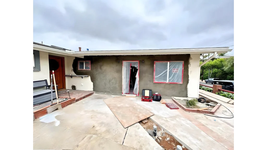 A single-story residential home under renovation showing fire damage, with charred areas around windows and doorways. The beige stucco exterior has visible burn marks, and windows are covered with red-taped protective plastic sheeting. The concrete driveway and front entrance area show construction materials and debris, indicating active restoration work.