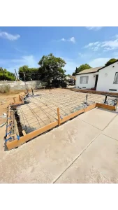 A construction site showing a concrete foundation slab with rebar reinforcement grid laid out in preparation for pouring. Wooden forms frame the perimeter of the foundation, with a white residential house visible in the background under a blue sky with scattered clouds. The site appears to be in a residential neighborhood with trees and fencing visible in the surrounding area.