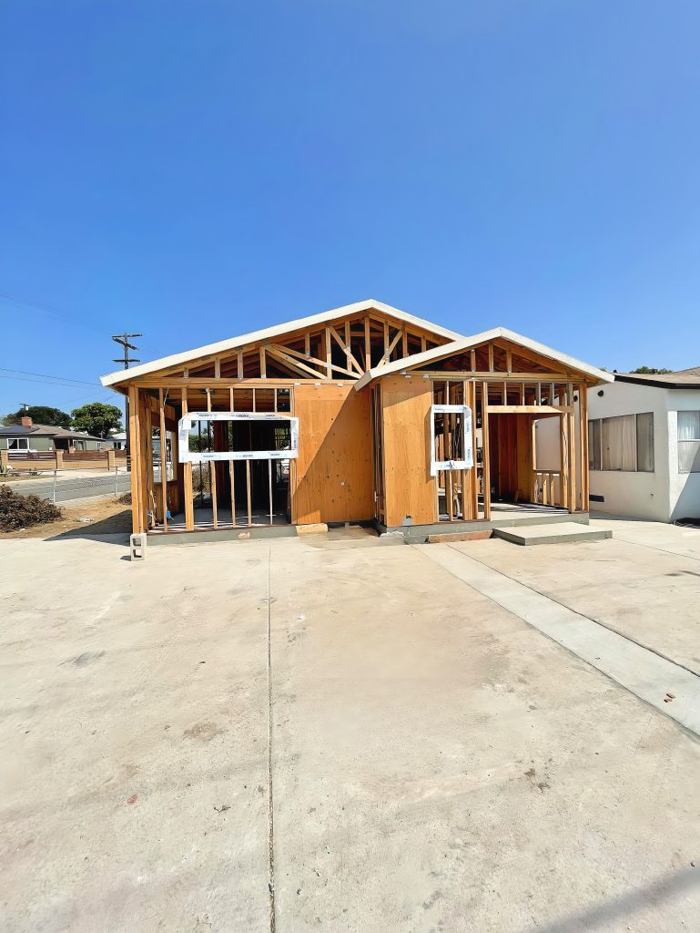 A house under construction showing exposed wood framing and roof trusses, with window and door openings visible in the structure. The building sits on a concrete foundation slab in what appears to be a residential neighborhood. An existing white stucco house is partially visible on the right side, with power lines and other homes in the background under a clear blue sky.