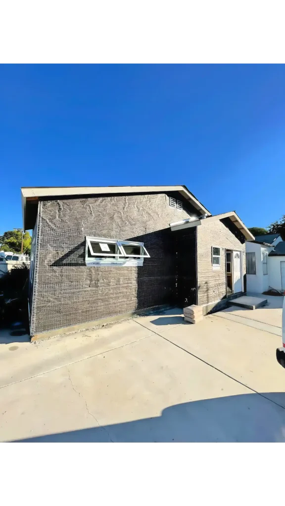 A modern two-story residential home featuring a distinctive angular design with mixed brick facades in black and tan colors. The contemporary architecture includes white-framed awning windows on the upper level and a clean concrete driveway. The house is photographed under a clear blue sky with neighboring homes visible in the background.