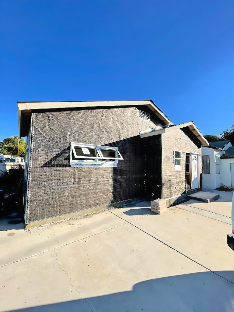 A modern two-story residential building under construction featuring a distinctive facade with light-colored brick on one side and dark charcoal or black brick on the other. The home has awning-style windows on the upper level and a concrete driveway leading to the entrance. The architectural design showcases clean lines and a contemporary aesthetic with a flat or low-slope roof against a clear blue sky.