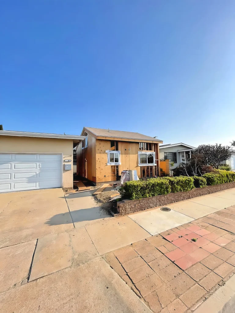 A residential property featuring a single-story house with natural wood siding and white-trimmed windows, attached to a cream-colored garage with white paneled door. The front yard has a brown brick retaining wall with neatly trimmed hedges, and the driveway consists of mixed concrete and terracotta-colored pavers. Neighboring houses are visible on either side under a clear blue sky.