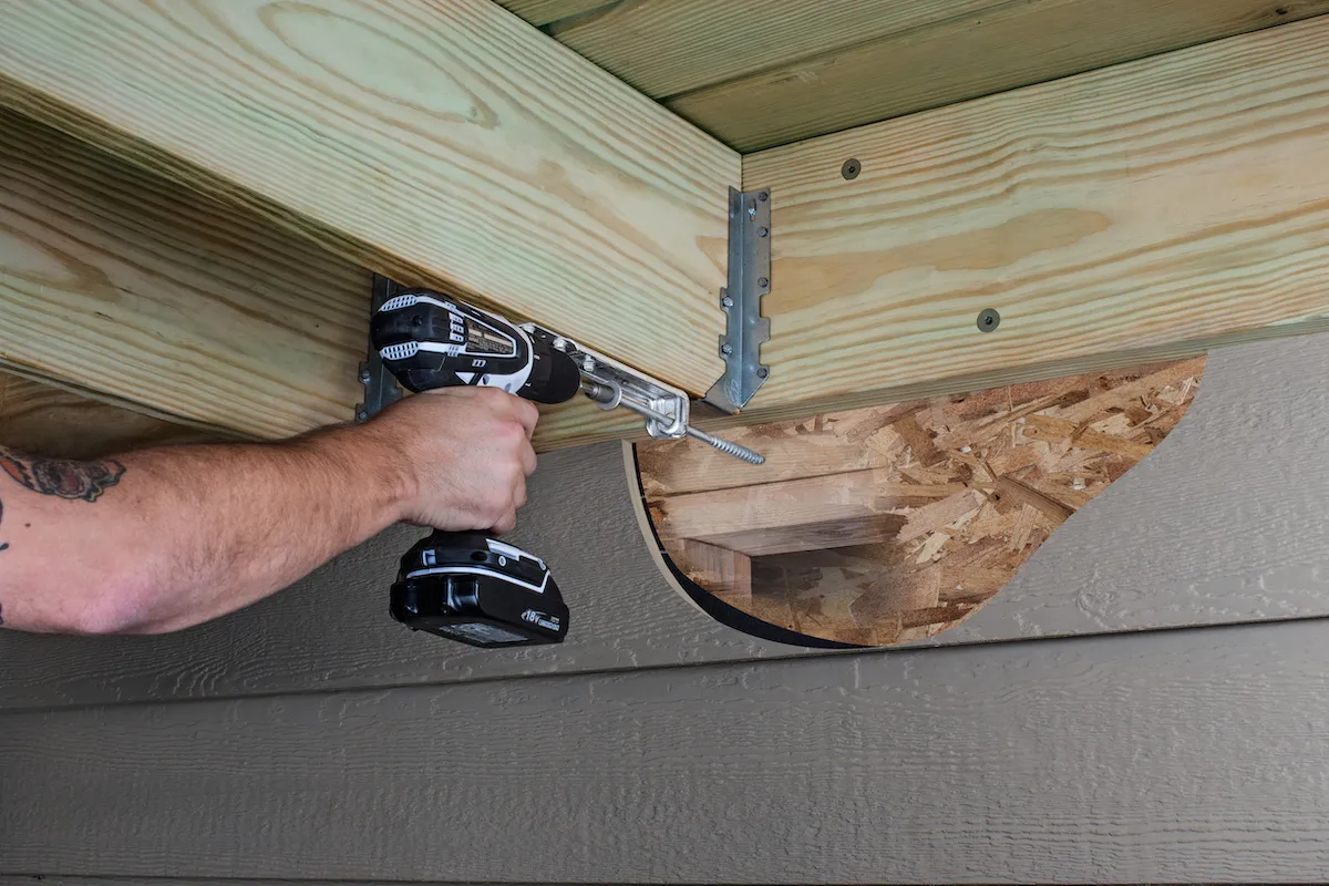 A person using a black power drill to install or secure wooden joists or beams in what appears to be a ceiling or overhead structure. Metal joist hangers are visible connecting the light-colored wooden boards, and there's a cable running through the framing. The exterior shows gray-colored siding beneath the wooden framework.