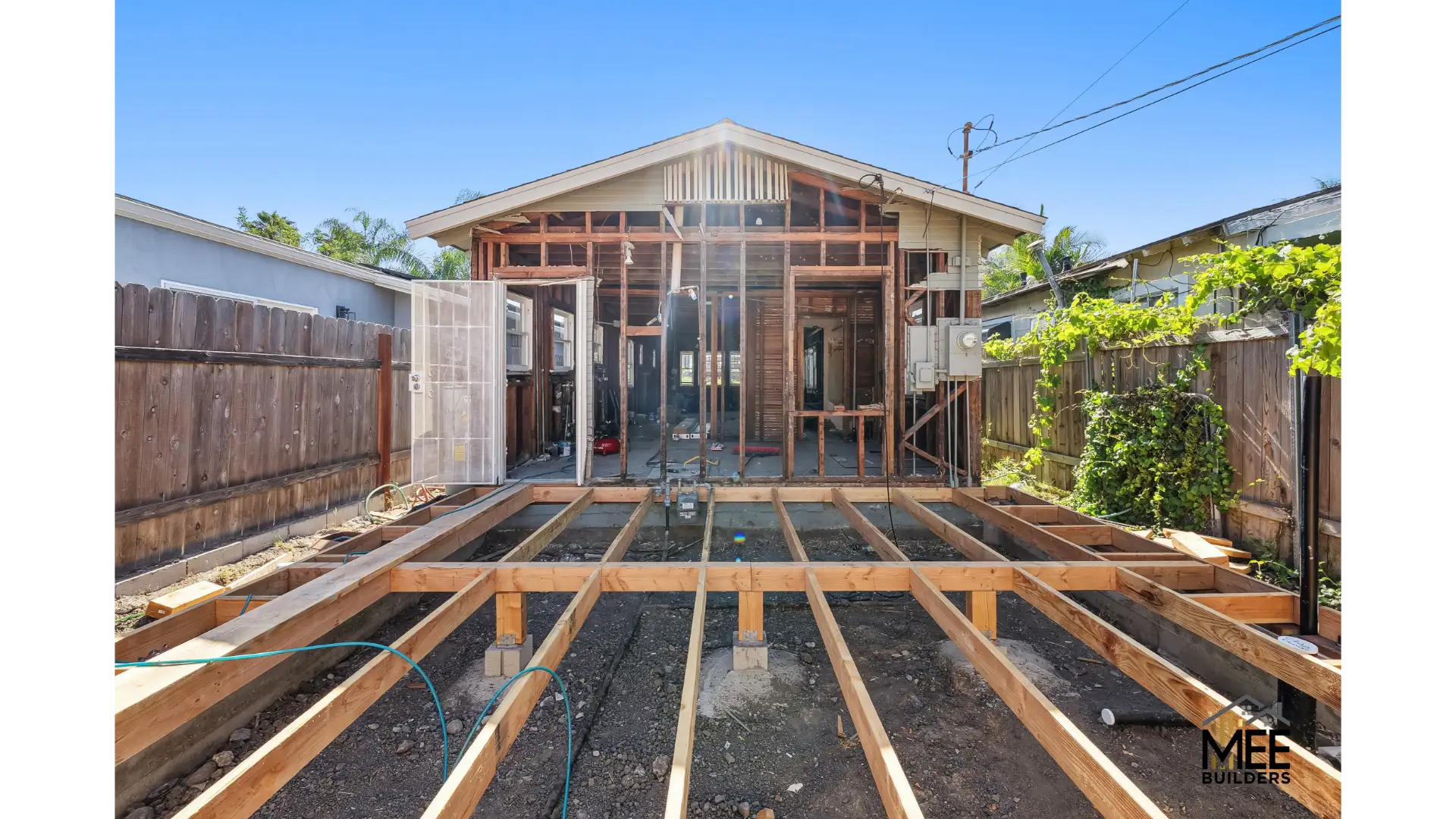 A residential home renovation in progress showing exposed floor joists made of lumber laid over prepared ground in the foreground, with the main house structure visible behind featuring wood framing and large window openings. The construction site is enclosed by wooden privacy fencing on both sides, with neighboring houses visible in the background under a clear blue sky. The outdoor renovation appears to be a room addition or deck construction project.