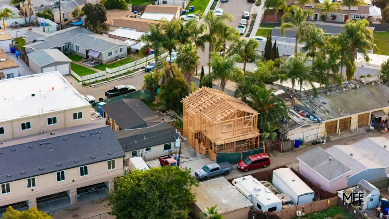 Aerial view of a residential neighborhood showing a two-story house under construction with exposed wooden framing and roof trusses. The structure stands prominently among completed single-story homes with various roof types, surrounded by palm trees and parked vehicles. The construction site is located on a densely packed suburban street with driveways and neighboring properties visible in all directions.