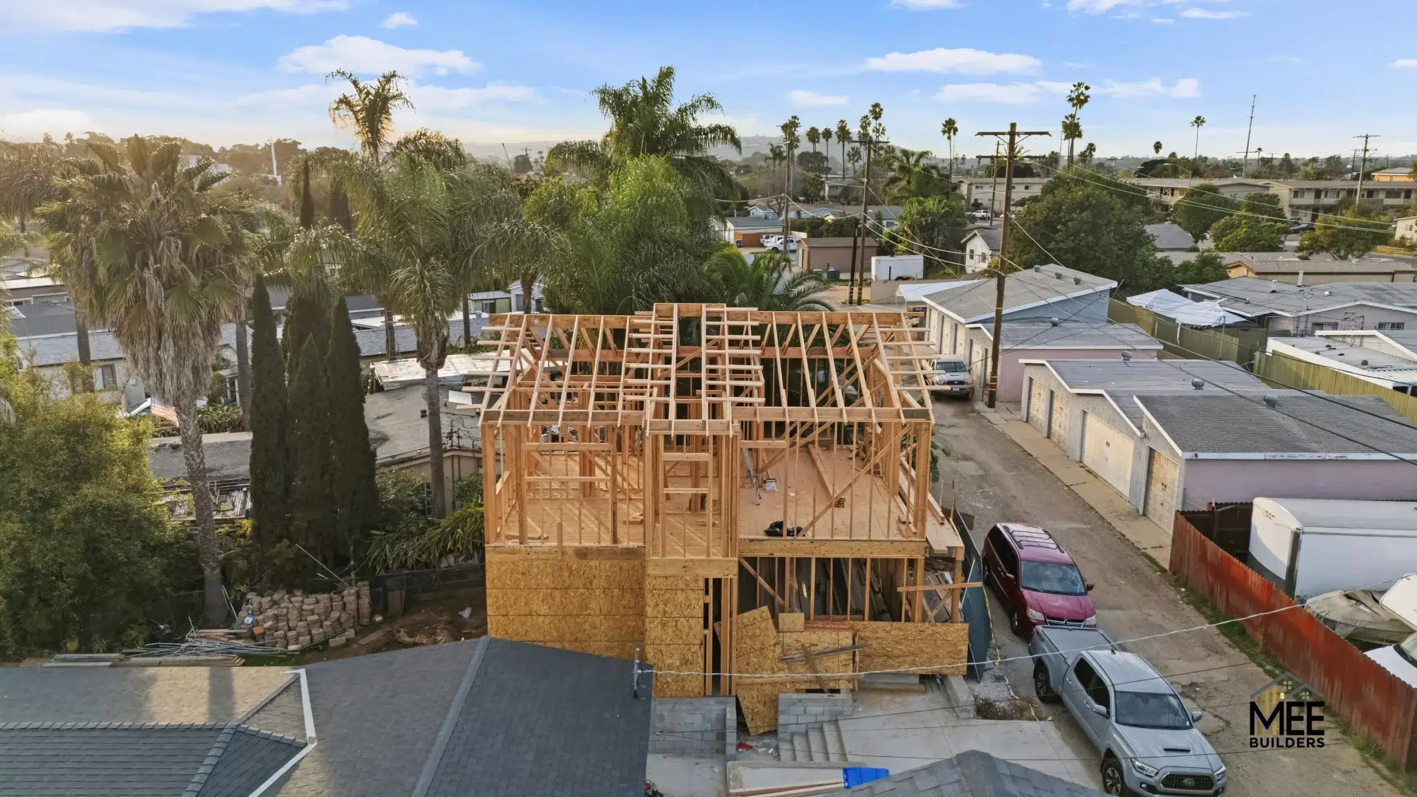 Aerial view of a two-story residential construction site showing the wooden frame and roof structure in progress. The house is surrounded by established neighborhood homes with palm trees and various vegetation visible throughout the suburban setting. Two vehicles are parked on the property, and the skeletal framework reveals the building's layout during the framing phase of construction.