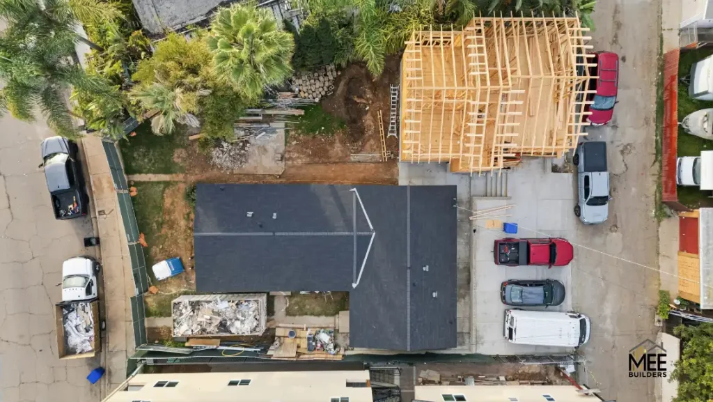 An aerial view of a residential construction site showing a dark gray flat roof with white line markings in the center, surrounded by wooden framing for a new structure. Several vehicles including trucks and cars are parked on the concrete driveway and street. Tropical vegetation and neighboring properties are visible around the construction area.