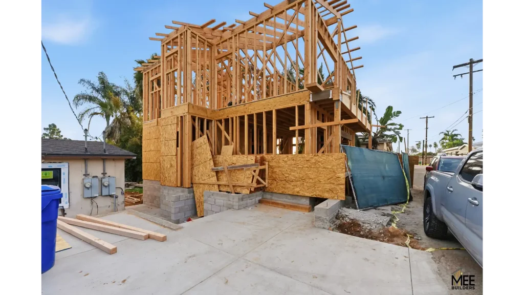 A two-story residential structure under construction showing exposed wooden framing and roof trusses. The building sits on a concrete foundation with cinder block walls, with plywood sheathing installed on the lower level. The construction site includes utility meters on an existing white structure to the left, palm trees in the background, and construction materials scattered around the concrete driveway.
