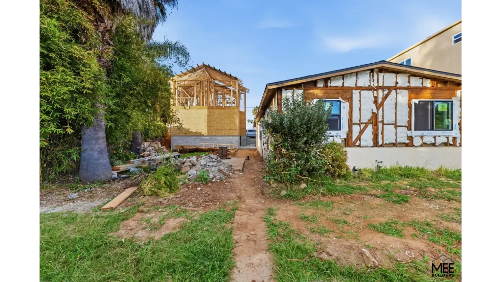 A residential construction site showing two houses in different stages of renovation. The house on the left has exposed wooden framing and is under active construction, while the house on the right displays exposed timber frame structure with white infill panels. The foreground shows a dirt pathway through patchy grass, with construction materials scattered around and mature trees providing greenery to the left side of the property.