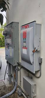 Two gray electrical utility boxes mounted on a pole outdoors, with the right box displaying red warning text or labels on its front panel. The boxes appear weathered and are surrounded by vegetation, with conduit connections visible at their bases. A white electrical component or meter is partially visible at the bottom of the frame.