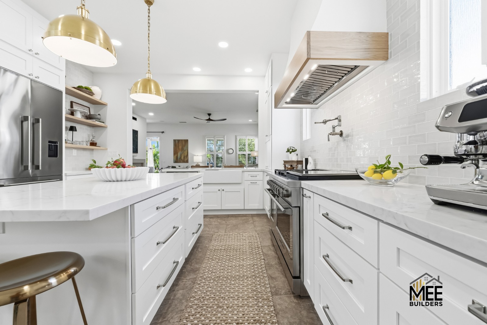 A modern white kitchen featuring a galley-style layout with white shaker cabinets, quartz countertops, and gold pendant lights. The space includes stainless steel appliances, white subway tile backsplash, a custom wood-paneled range hood, and a kitchen island with bar seating. The design showcases a clean, bright aesthetic with natural wood accents and views into adjacent living spaces.