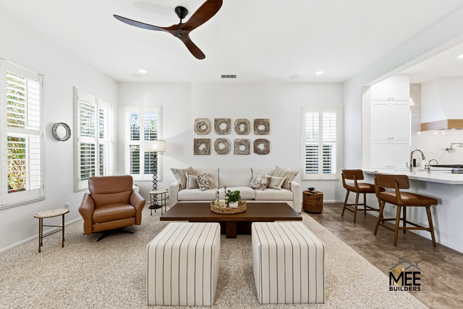 A bright, modern open-concept living space featuring a white color palette with warm wood accents. The room includes a beige sofa with decorative pillows, a brown leather armchair, striped ottomans, and a dark wood coffee table, all beneath a contemporary ceiling fan. White plantation shutters line the windows, and the space flows into a white kitchen area with wooden bar stools and decorative wall art.