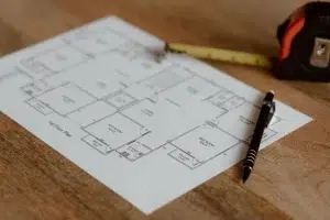 Architectural floor plan blueprint laid on a wooden surface with a black pen and yellow tape measure placed on top. The technical drawing shows the layout of a building with multiple rooms and labeled spaces. The image represents construction or home renovation planning materials.