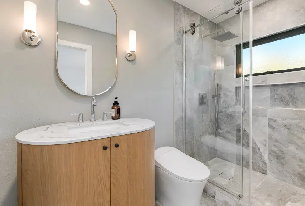 A modern bathroom featuring a curved wooden vanity with white marble countertop and chrome fixtures, accompanied by a U-shaped mirror with wall sconces. The space includes a white toilet and glass-enclosed shower with gray textured tiles, while natural light enters through a window visible in the reflection.