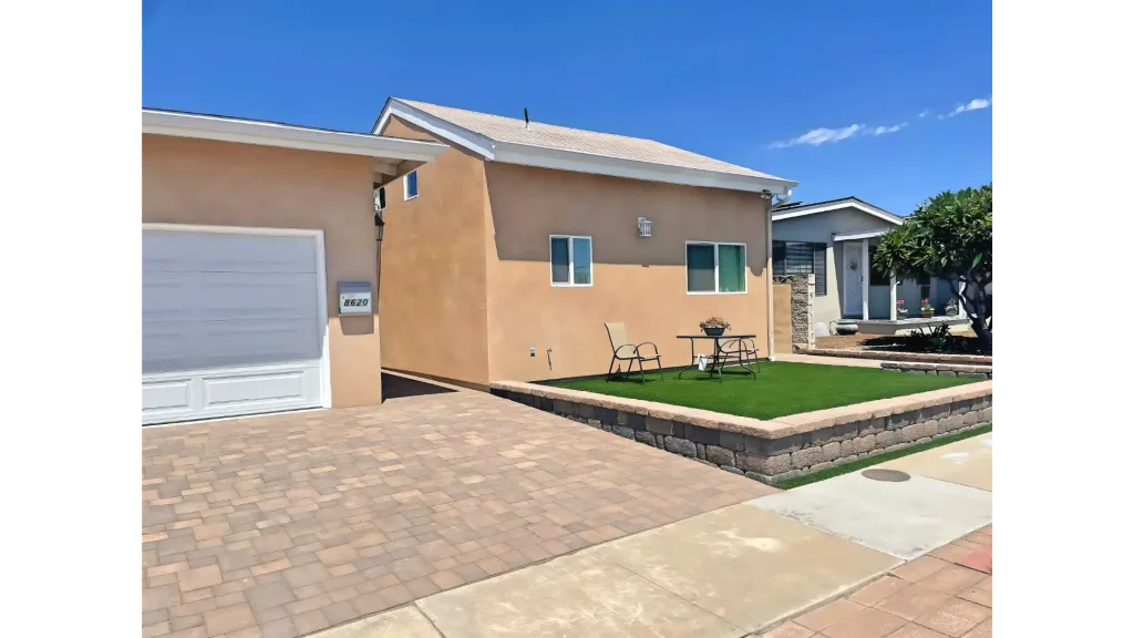 A suburban residential property featuring a peach-colored stucco house with white trim and a white garage door. The front yard showcases a raised lawn area with artificial turf bordered by decorative stone pavers, with patio furniture placed on the grass. A paver driveway leads to the garage, and neighboring houses are visible in the background under a clear blue sky.
