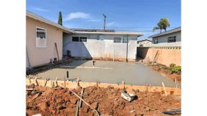 A freshly poured concrete slab in a residential backyard, surrounded by wooden formwork and excavated red-brown soil. The smooth gray concrete surface appears level and is in the process of curing, with stakes and forms still in place. A single-story beige house with windows is visible in the background, along with neighboring properties and palm trees under a clear blue sky.