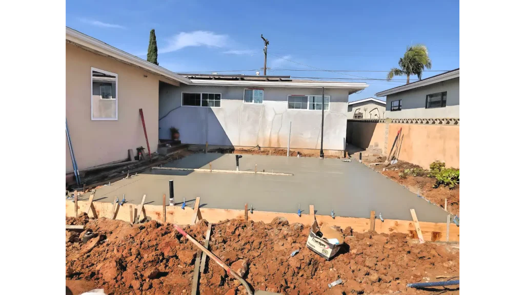 A freshly poured concrete slab in a residential backyard, surrounded by wooden forms and disturbed reddish-brown soil. The smooth gray concrete surface extends between a beige house on the left and a neighboring property visible behind a white fence. Construction materials and equipment are scattered around the work area under a clear blue sky with palm trees visible in the background.