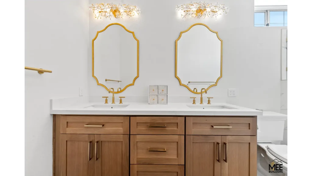 A modern bathroom vanity featuring a white marble countertop with dual sinks and gold fixtures. Two ornate gold-framed mirrors hang above the sinks, complemented by elegant crystal and gold light fixtures. The cabinetry is natural wood with a warm honey tone and brass hardware, creating a luxurious contemporary design.