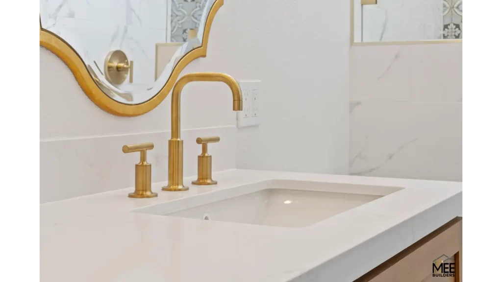 A luxurious bathroom vanity featuring a white countertop with an undermount sink and elegant brushed gold three-piece faucet set. The space is adorned with a decorative gold-framed mirror with curved edges mounted above white marble tile backsplash. The design combines classic sophistication with modern elements, creating an upscale bathroom aesthetic.