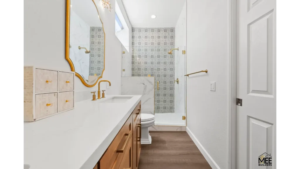 A modern bathroom featuring white quartz countertops, warm wood cabinetry, and elegant gold fixtures including a curved mirror frame and faucet. The shower area showcases decorative patterned tile work in gray and white, enclosed by frameless glass. The space has light gray walls, wood-look flooring, and gold hardware throughout, creating a sophisticated blend of traditional and contemporary design.