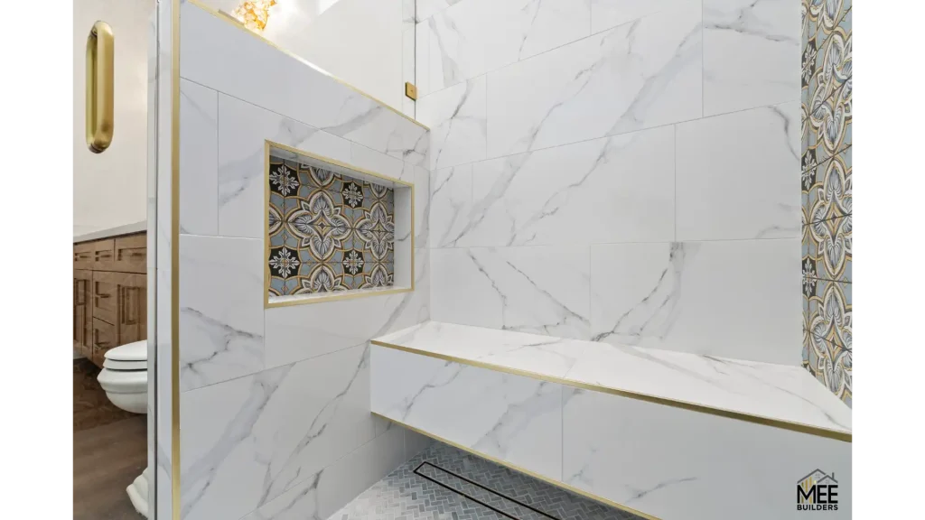 A luxurious modern bathroom featuring floor-to-ceiling white marble tiles with grey veining, accented by elegant gold trim. The space includes a built-in shower bench with gold edging, a decorative recessed niche with ornate patterned tiles in blue, gold and grey tones, and a glass shower enclosure with gold hardware. The design combines classic elegance with contemporary minimalism, showcasing high-end finishes throughout.