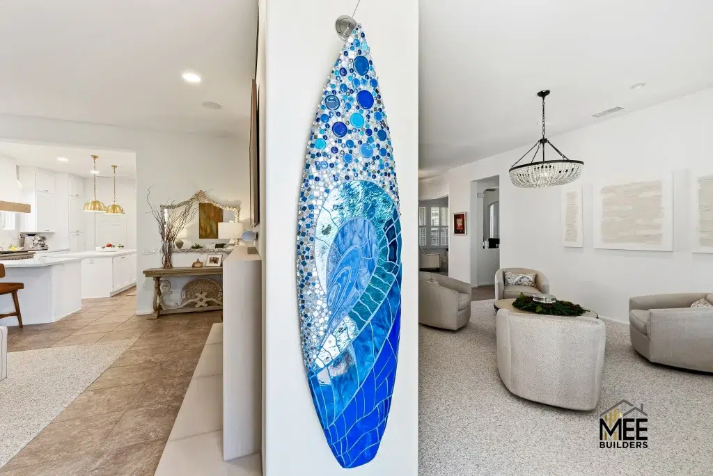 A decorative surfboard with blue mosaic tile artwork hangs vertically on a white wall between an open-plan kitchen and living room. The surfboard features various shades of blue tiles creating an ocean wave pattern, with circular bubble-like designs at the top transitioning to geometric patterns below. The modern coastal home interior showcases white cabinetry, beige furniture, and neutral tones throughout the space.