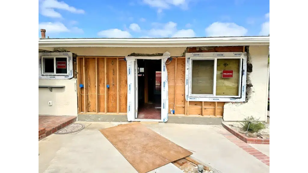 A single-story house undergoing major renovation with its exterior wall torn open, revealing exposed wooden framing and interior spaces. Three new white-framed windows are installed, two of which still have protective labels, while the stucco exterior shows significant damage around the openings. A piece of cardboard lies on the concrete driveway in front of the construction zone.